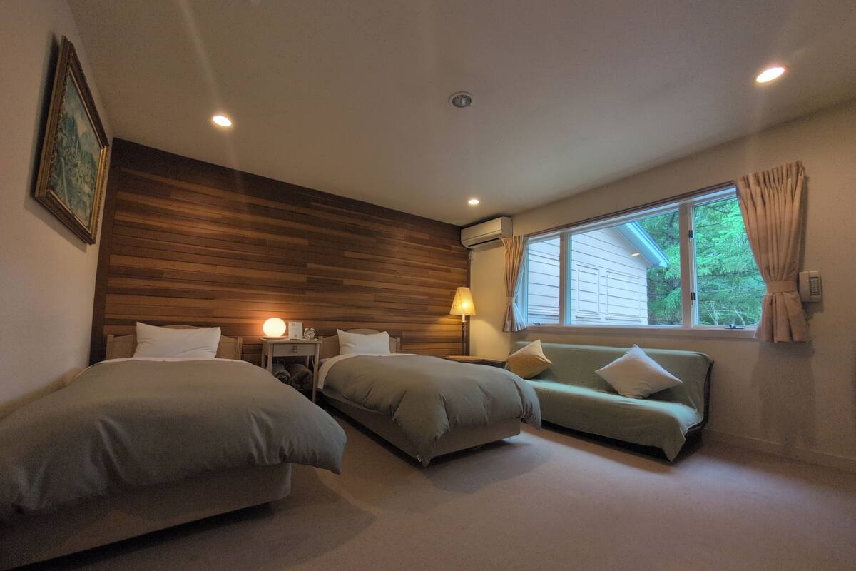 A hotel room with two beds, a sofa, a lamp, an air conditioning unit, and a window with curtains.
