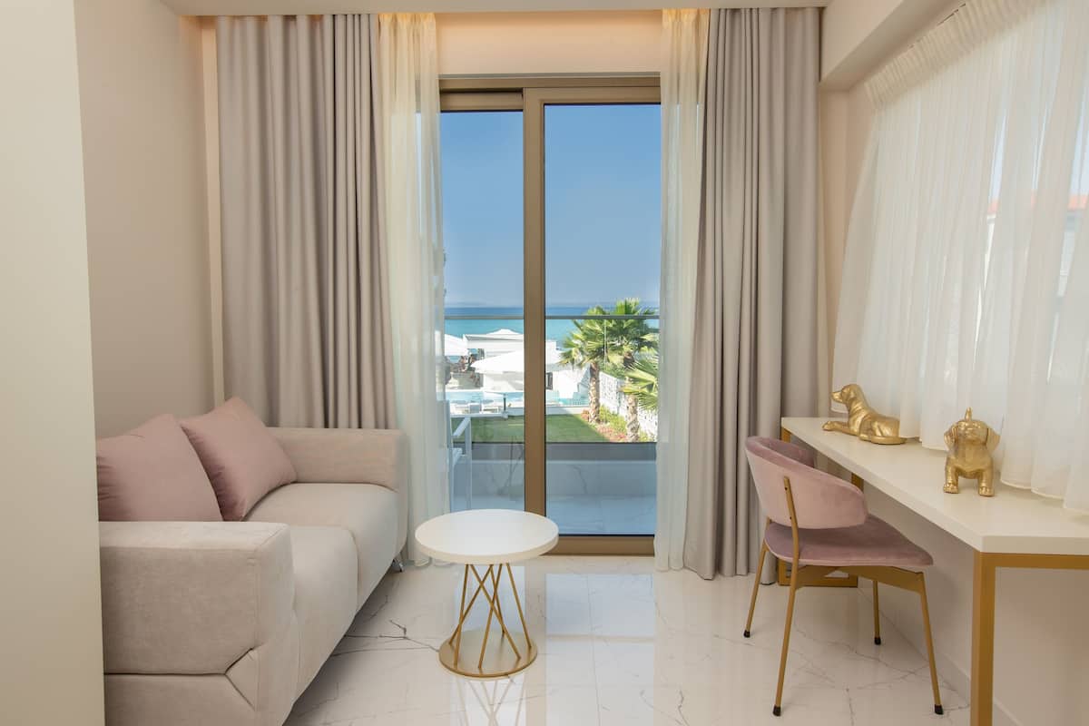 Una sala de estar moderna con un sofá, una mesa redonda y un comedor con una silla. Un amplio ventanal corredero da a un balcón con vistas al mar y palmeras.