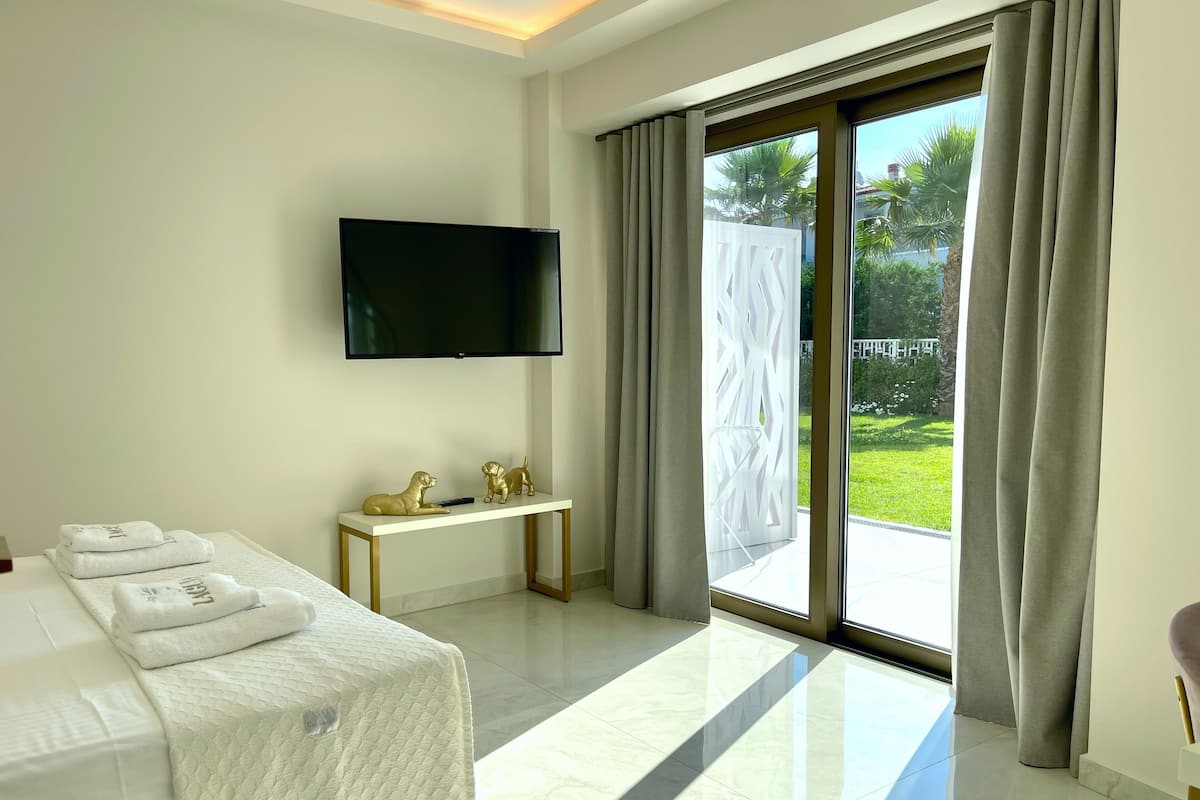Habitación de hotel con una cama, un televisor de pantalla plana montado en la pared, una mesita con toallas y vista a un jardín por una puerta corrediza de vidrio.