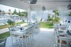 Un restaurant avec des tables et des chaises blanches, une terrasse et une vue sur une piscine et un espace verdoyant.