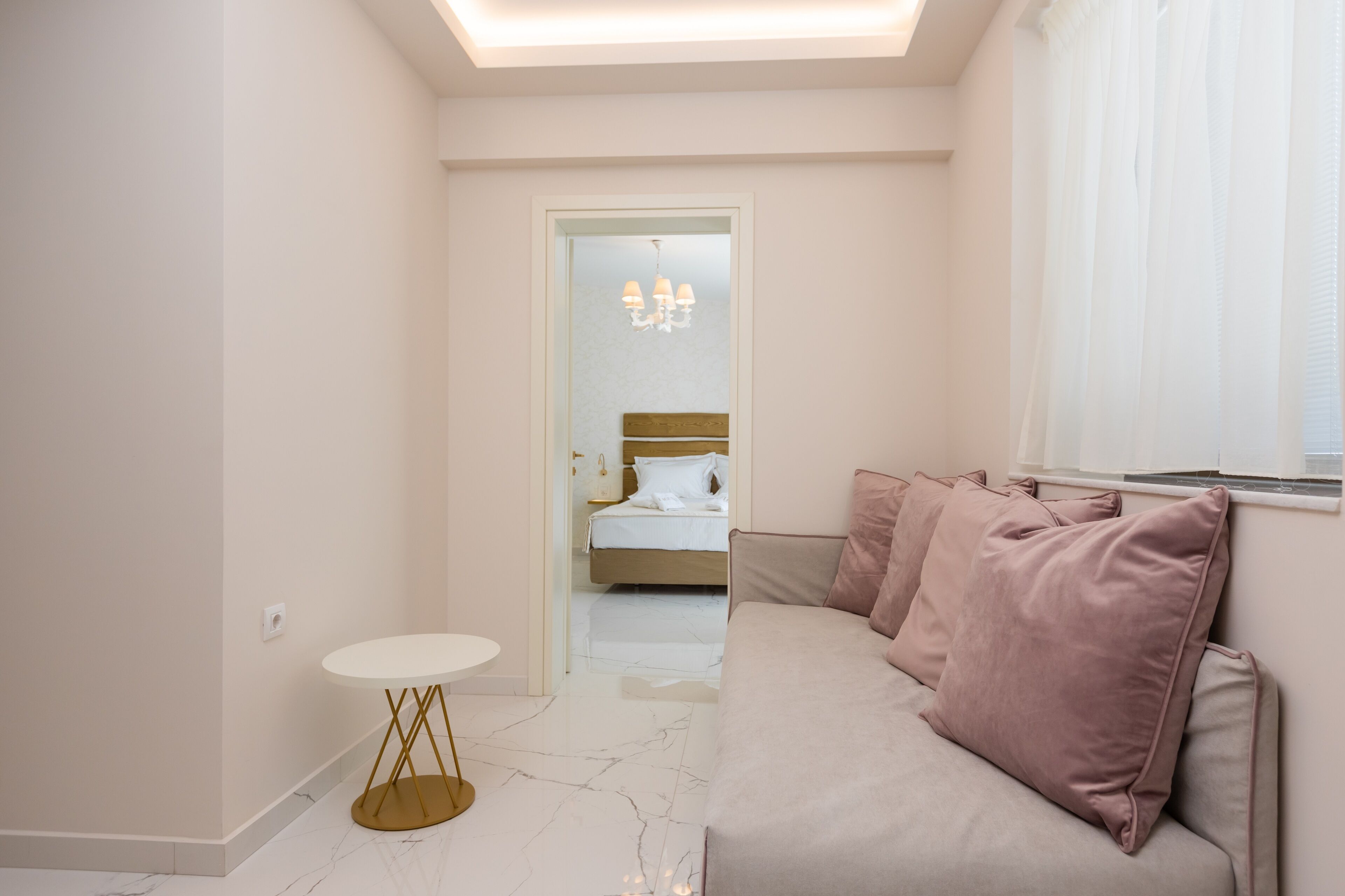 A modern hotel room with a marble floor, a round table with a gold base, a beige sofa with pink cushions, and a bed with white linens.