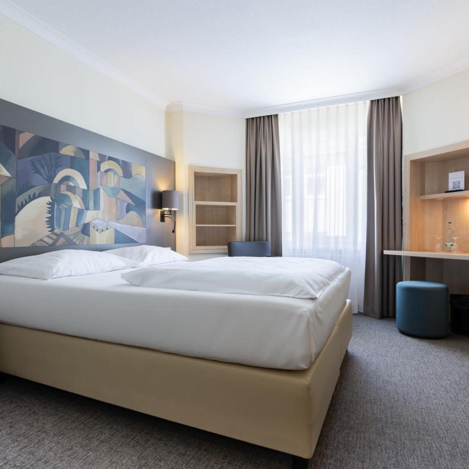 A hotel room with a large bed, a desk, a chair, a bookshelf, and a painting on the wall.