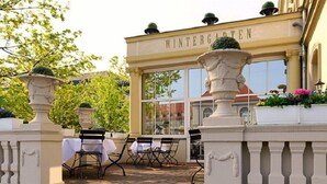 A building entrance with the sign "WINTERGARTEN" above a glass door, outdoor seating with tables and chairs, and decorative urns on the steps. - Kaiserin Augusta (Weimar)