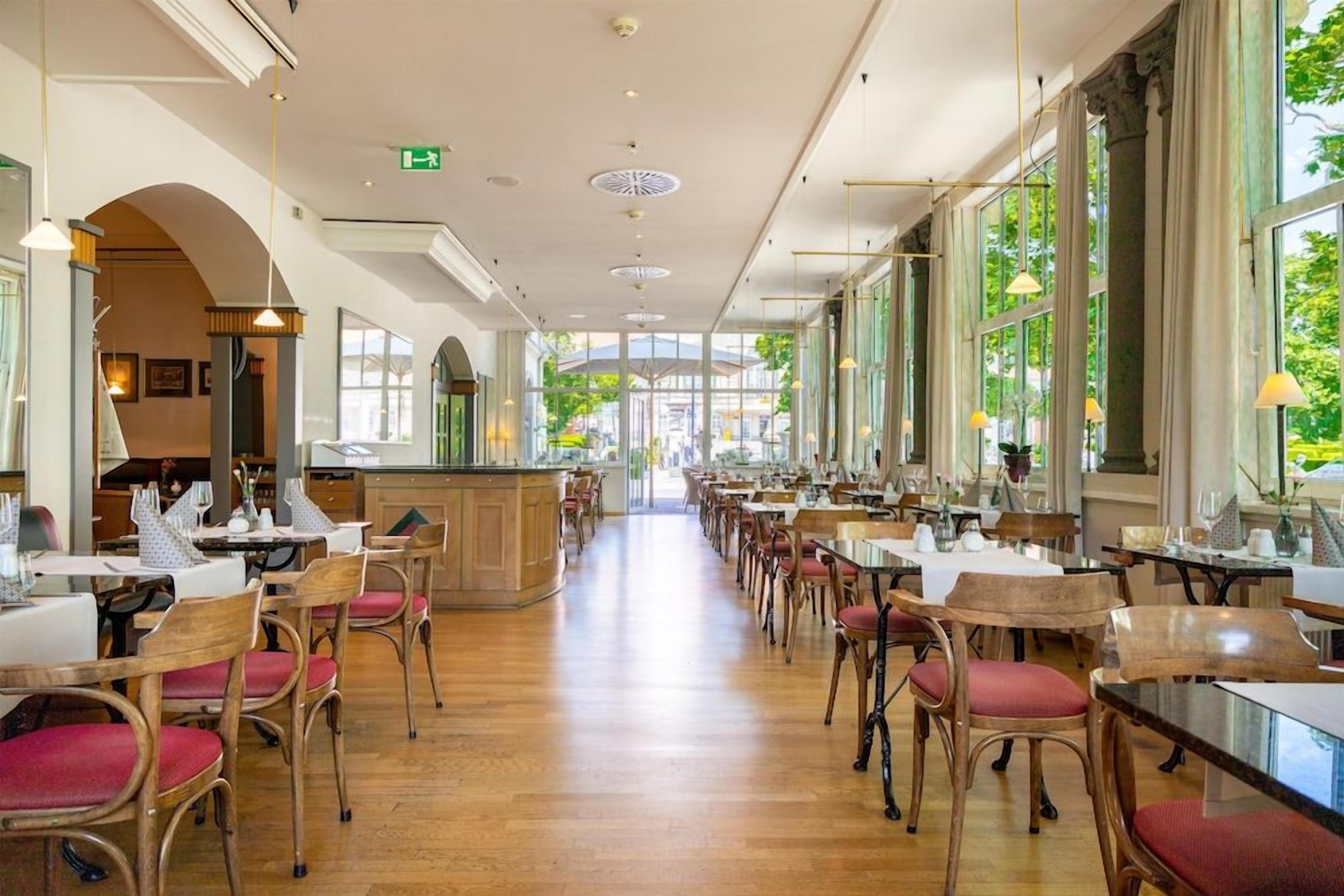 A spacious restaurant with wooden flooring, red cushioned chairs, and large windows allowing natural light.