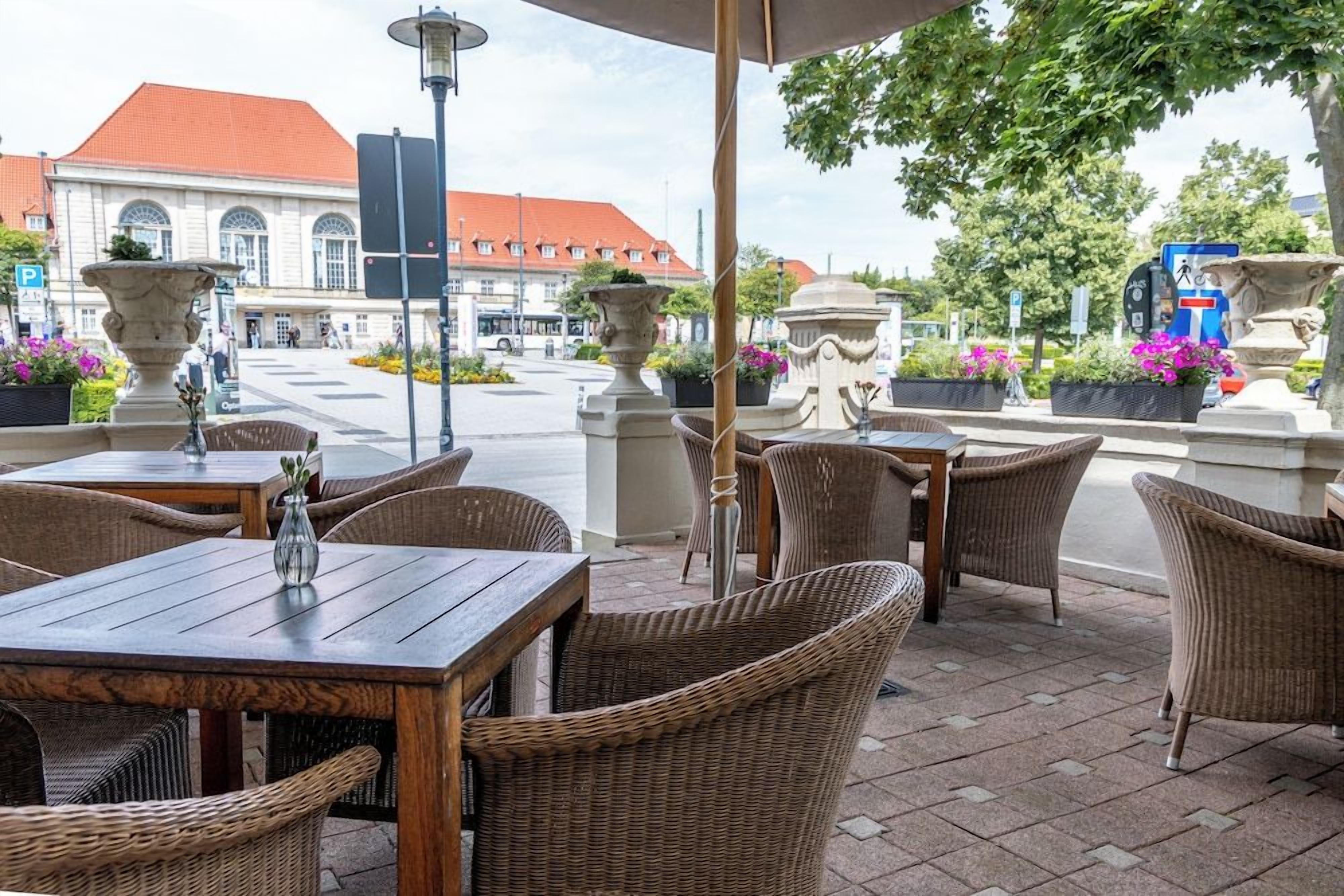 Ein Sitzbereich im Freien mit Korbmöbeln und Steinpflanzgefäßen, ein Straßenbild mit einem Gebäude im Hintergrund und ein blaues Schild mit Fahrradsymbol.