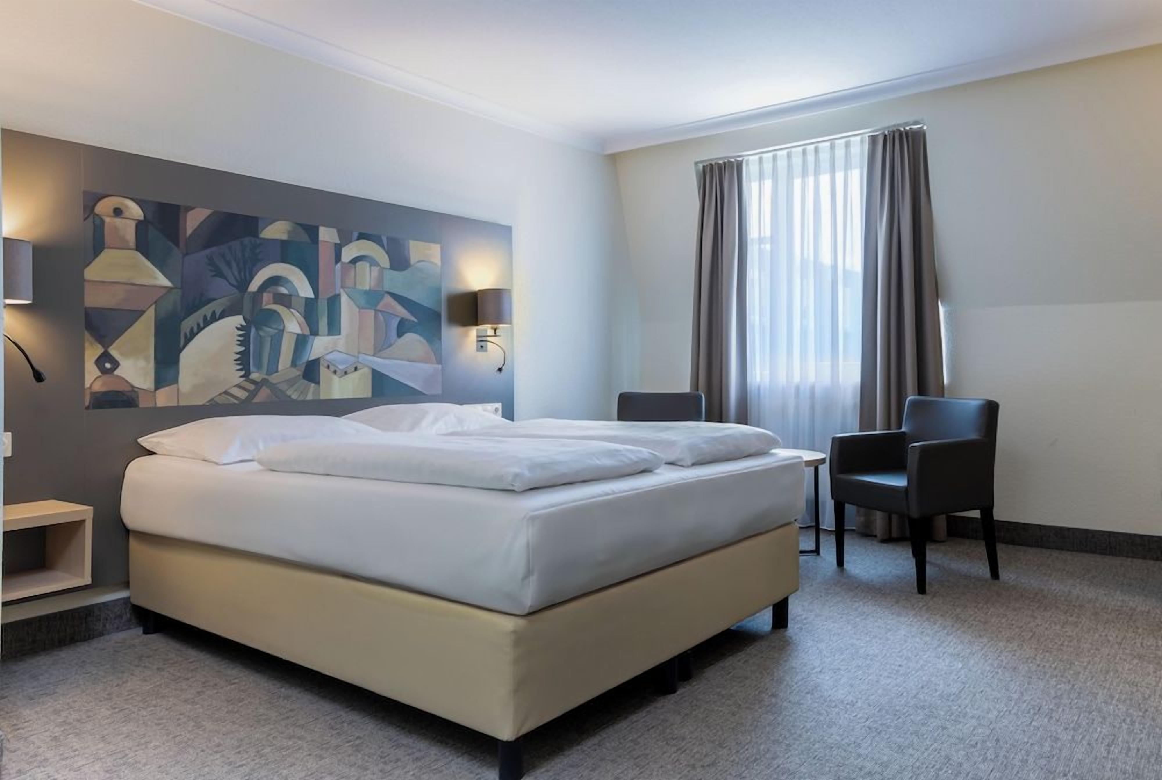 Quarto de hotel com cama, duas cadeiras, escrivaninha, abajur e um quadro na parede.
