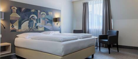 Quarto de hotel com cama, duas cadeiras, escrivaninha, abajur e um quadro na parede.