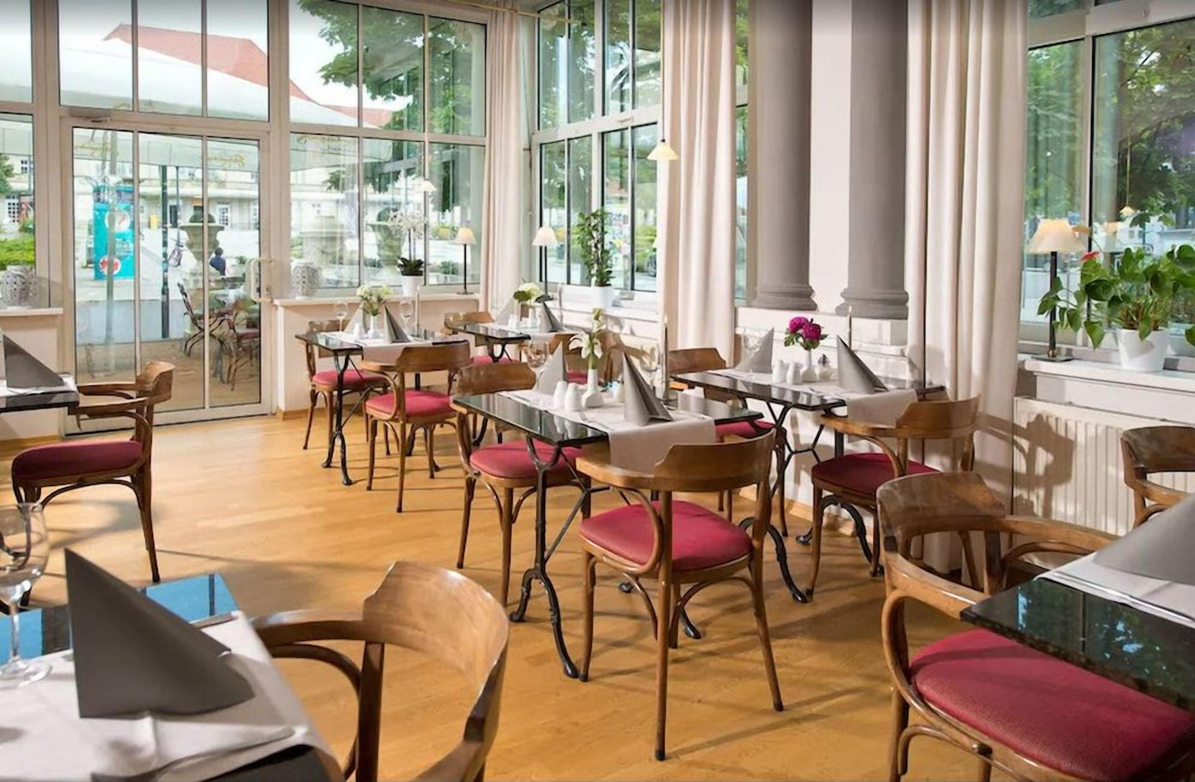 Ein Restaurant mit Holztischen und -stühlen, großen Fenstern und Blumentöpfen.