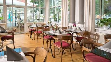 Ein Restaurant mit Holztischen und -stühlen, großen Fenstern und Blumentöpfen.