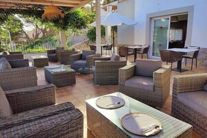 Un área de estar al aire libre con mobiliario de mimbre y una mesa de vidrio, rodeada por un muro blanco y una puerta de cristal.
