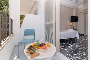 Habitación de hotel con cama, televisor montado en la pared, una mesita con fruta y un libro, y un balcón con una silla.