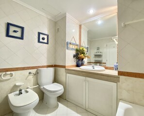 Un baño con inodoro, bidé y lavamanos. Hay cuadros enmarcados en la pared y un espejo sobre el lavamanos.