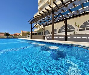 Una piscina con estructura de pérgola, agua azul cristalina y un edificio con ventanas de arco en el fondo.