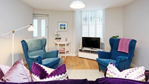A living room with two blue armchairs, a white TV stand, a floor lamp, and a window with blinds. - JOIVY Vibrant City Centre 2-bed Apt. (Edinburgh)