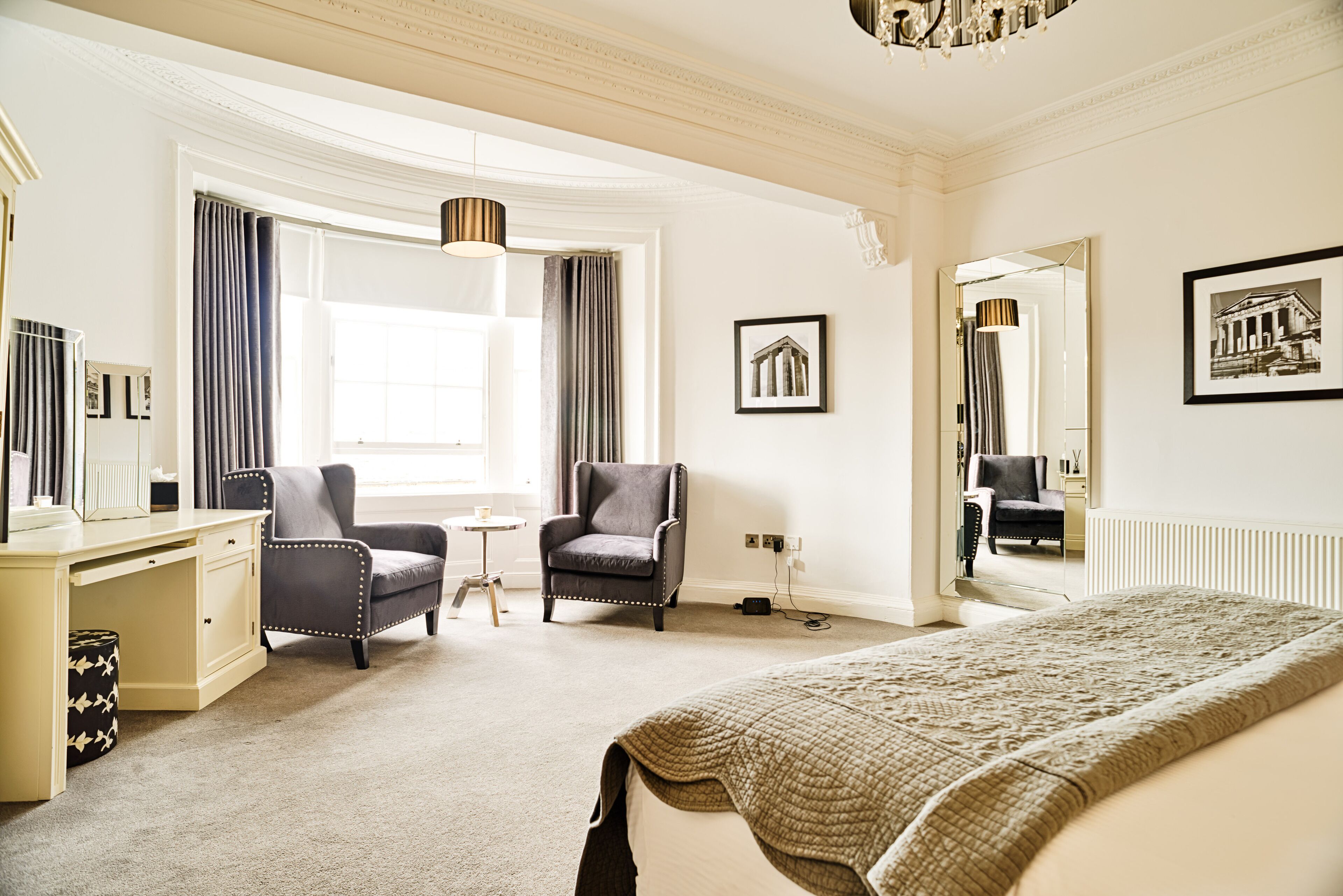 A hotel room with a large bed, a desk, two armchairs, a small table, and a large window with curtains.