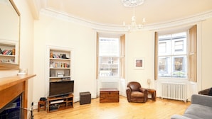 Un salón con piso de madera, chimenea, televisor, estantería, sillón de cuero y ventana con vista a edificios.