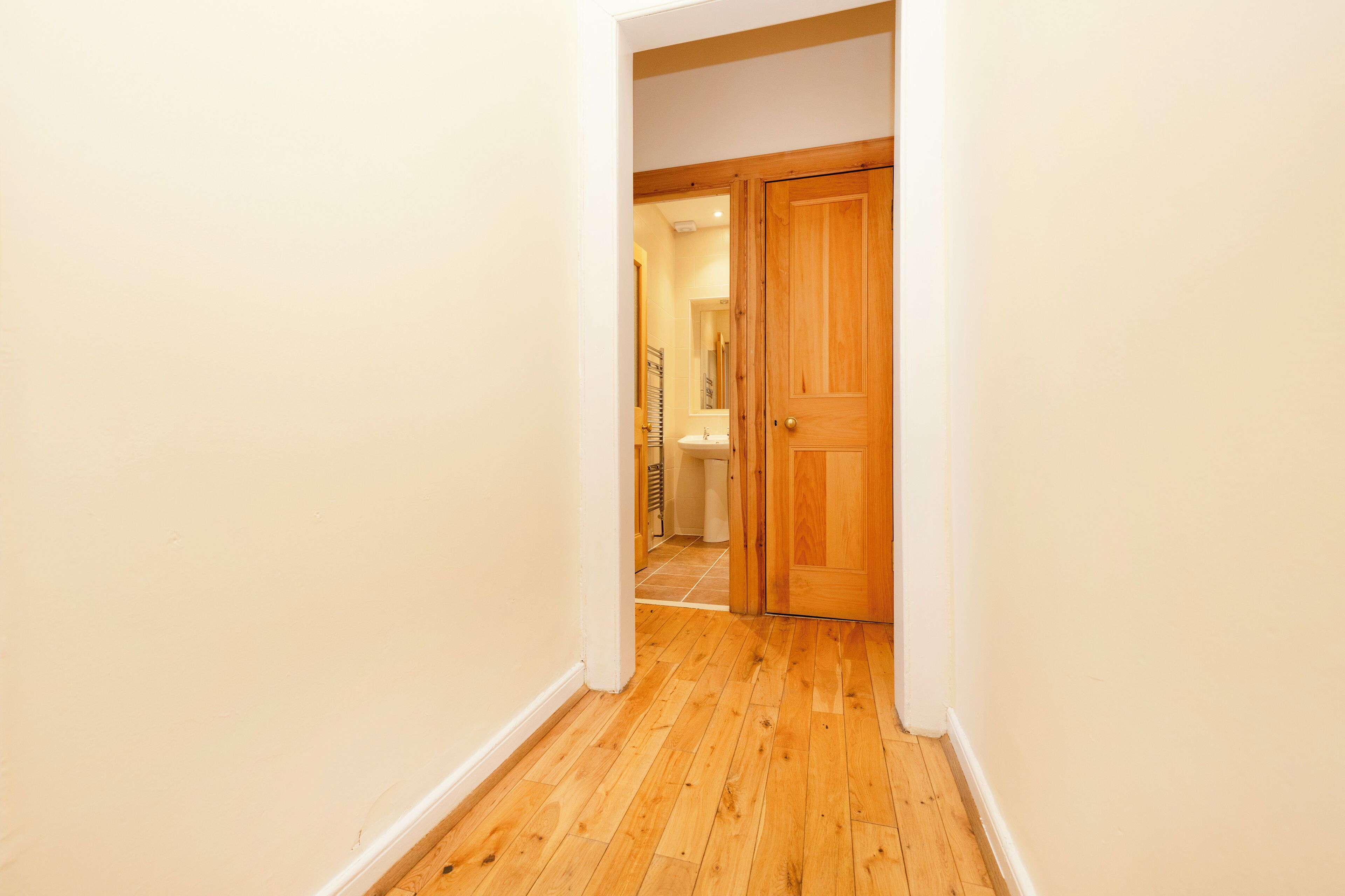Un couloir au sol en bois et aux murs blancs mène à une salle de bain équipée d’un lavabo et d’un miroir.