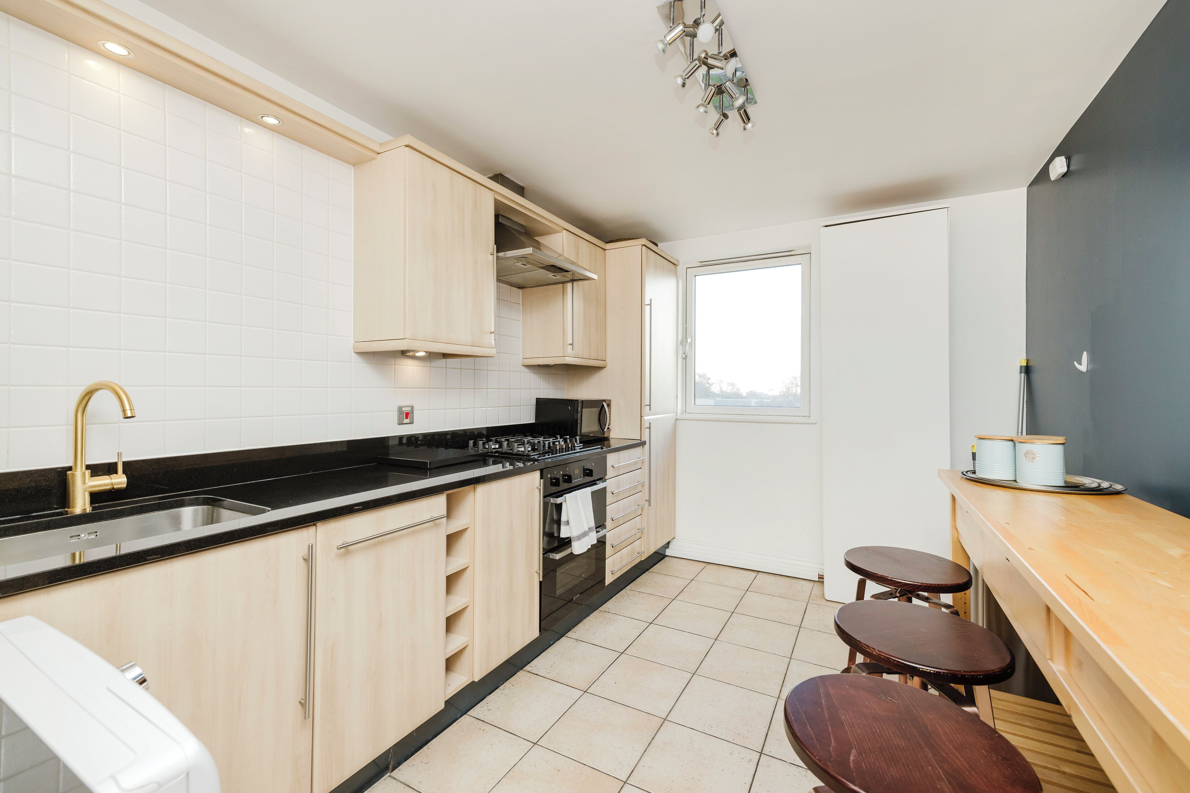 A modern kitchen with light wood cabinets, a black countertop, a stainless steel sink, and a built-in oven. There are two wooden stools and a window providing natural light.