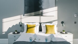 A modern bedroom with a large bed, two bedside tables, a wall-mounted lamp, and a painting.