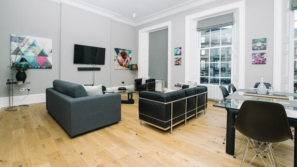 A modern living room with a grey sofa, a black sectional, and a glass coffee table. There are abstract paintings on the walls and a flat-screen TV mounted above the sofa.