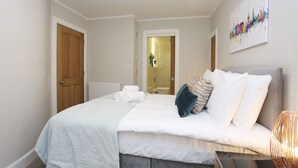 Una cama bien hecha con ropa de cama blanca y un cabecero gris, una puerta de madera a la izquierda, y un baño visible a través de una puerta abierta al fondo.