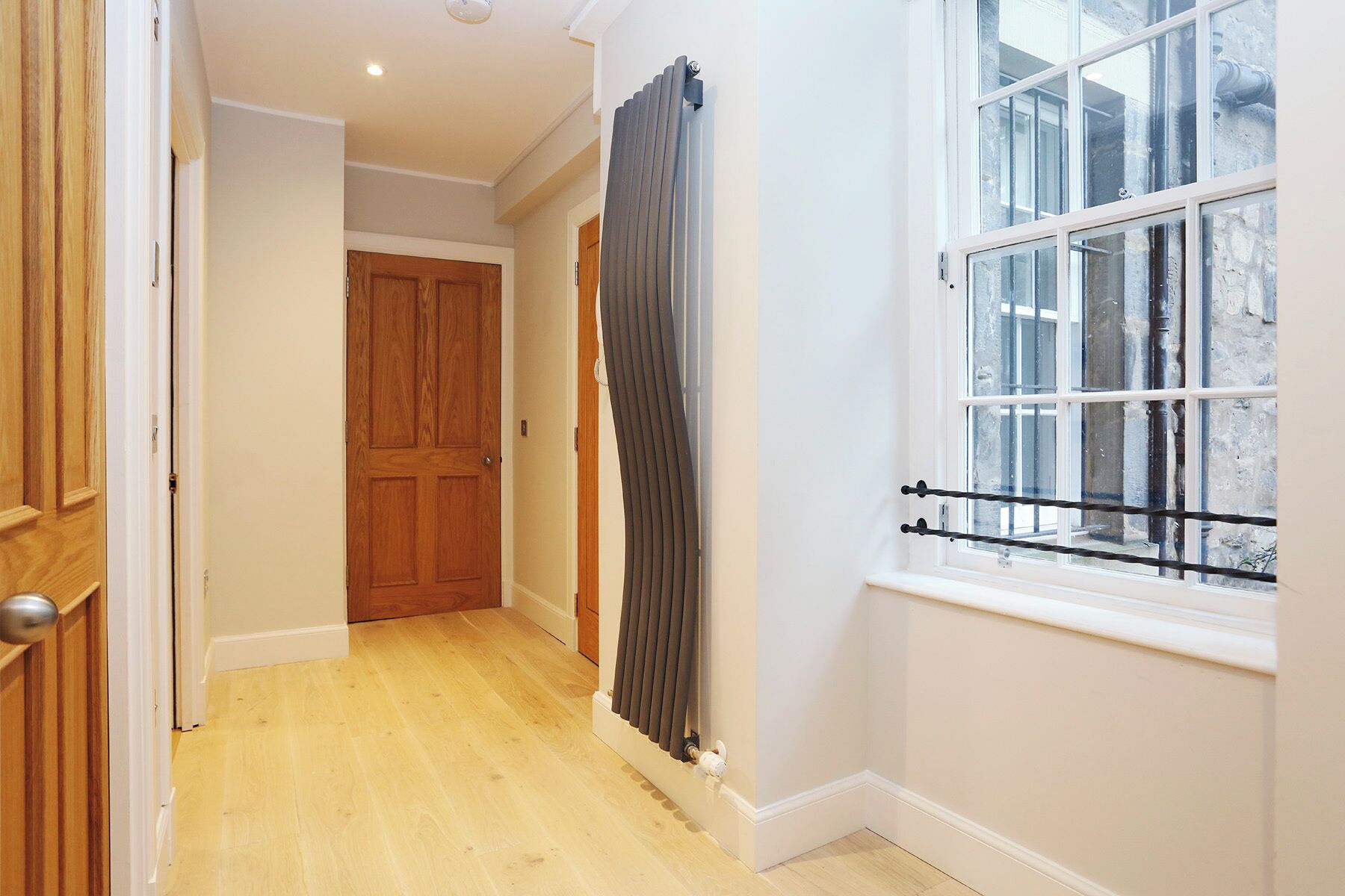 Un couloir avec un plancher en bois, une porte en bois, un radiateur et une fenêtre avec des stores.