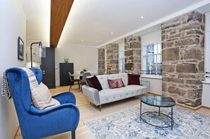 A modern living room with a blue armchair, a grey sofa with red cushions, a glass coffee table, and a patterned rug.