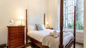 A four-poster bed with a white bedspread and two pillows, a wooden dresser with drawers, two bedside lamps, and a window with a view of greenery. - JOIVY Converted Flat in Historic Building in Desirable New Town (Edinburgh)