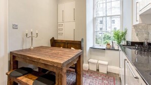 A kitchen with a wooden table, white cabinets, and a window with a view of greenery. - JOIVY Converted Flat in Historic Building in Desirable New Town (Edinburgh)