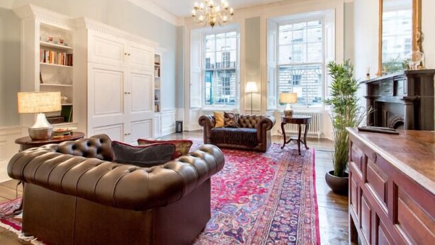A spacious living room with a large brown leather sofa, a patterned rug, and a view of the city through large windows. - JOIVY Converted Flat in Historic Building in Desirable New Town (Edinburgh)