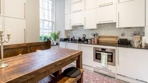 A modern kitchen with white cabinets, a wooden dining table, and a large window providing natural light. - JOIVY Converted Flat in Historic Building in Desirable New Town (Edinburgh)