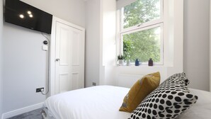 A bedroom with a bed, a window with a view of trees, a TV mounted on the wall, and a door. - JOIVY Stunning Ground-floor 2bedrm New Town Apt (Edinburgh)