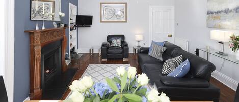 A living room with a fireplace, a black leather sofa, a glass coffee table, and a chandelier.