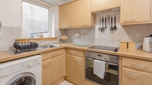 A compact kitchen with wooden cabinets, a washing machine, an oven, and a window providing natural light. - JOIVY Heart of The Royal Mile (Edinburgh)