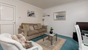 A living room with a beige sofa, a white armchair, a glass coffee table, a patterned rug, a ceiling light fixture, and a wall-mounted TV. - JOIVY Heart of The Royal Mile (Edinburgh)