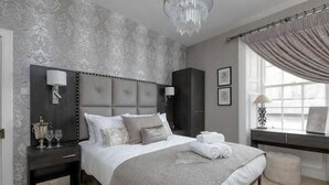 Un dormitorio con una cama grande, cabecera oscura, lámparas de noche, una mesita de noche y una ventana con cortinas.