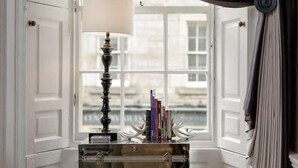 Una esquina de una habitación con una ventana, una lámpara y una mesita con libros.