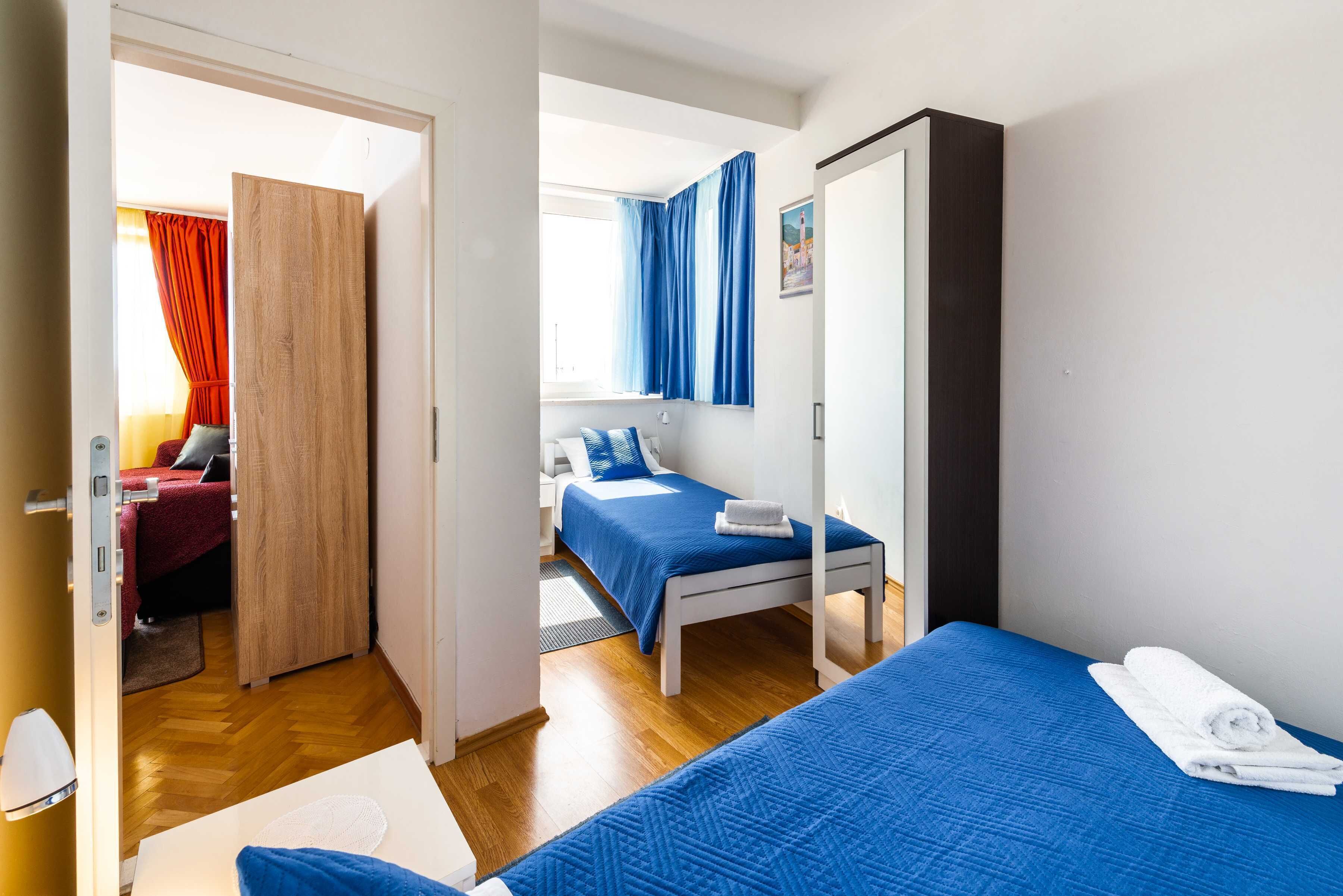 A hotel room with two beds, a wooden wardrobe, a desk with a lamp, and a view of another room with a red curtain.