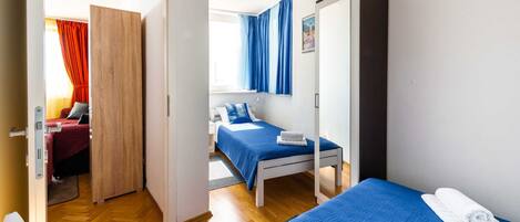 A hotel room with two beds, a wooden wardrobe, a desk with a lamp, and a view of another room with a red curtain.