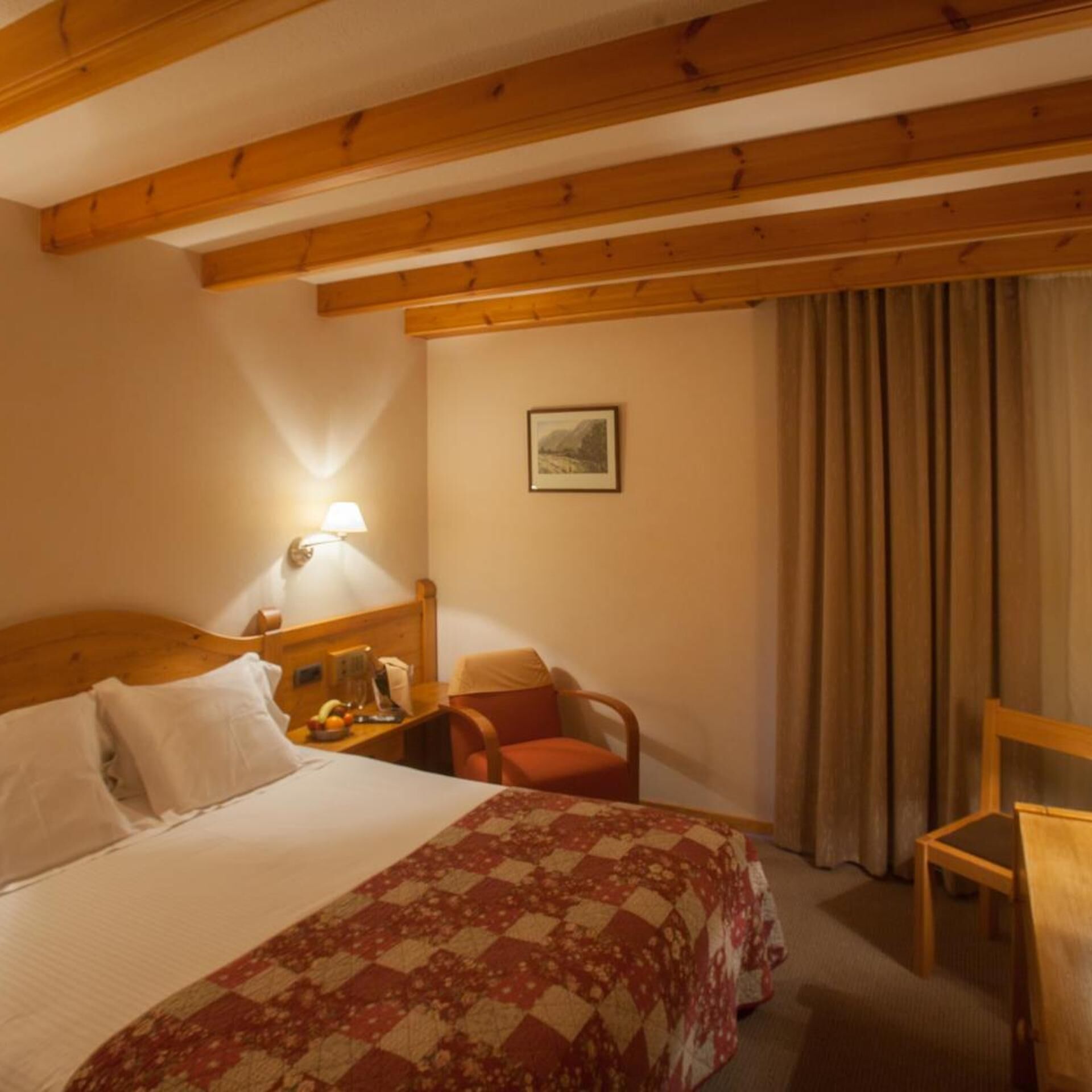 Habitación de hotel con cama, una silla, escritorio con lámpara, un cuadro en la pared y ventana con cortinas.