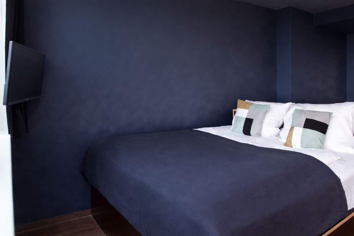 A modern hotel room with a dark blue wall, a large bed with white and patterned pillows, and a flat-screen TV mounted on the wall.