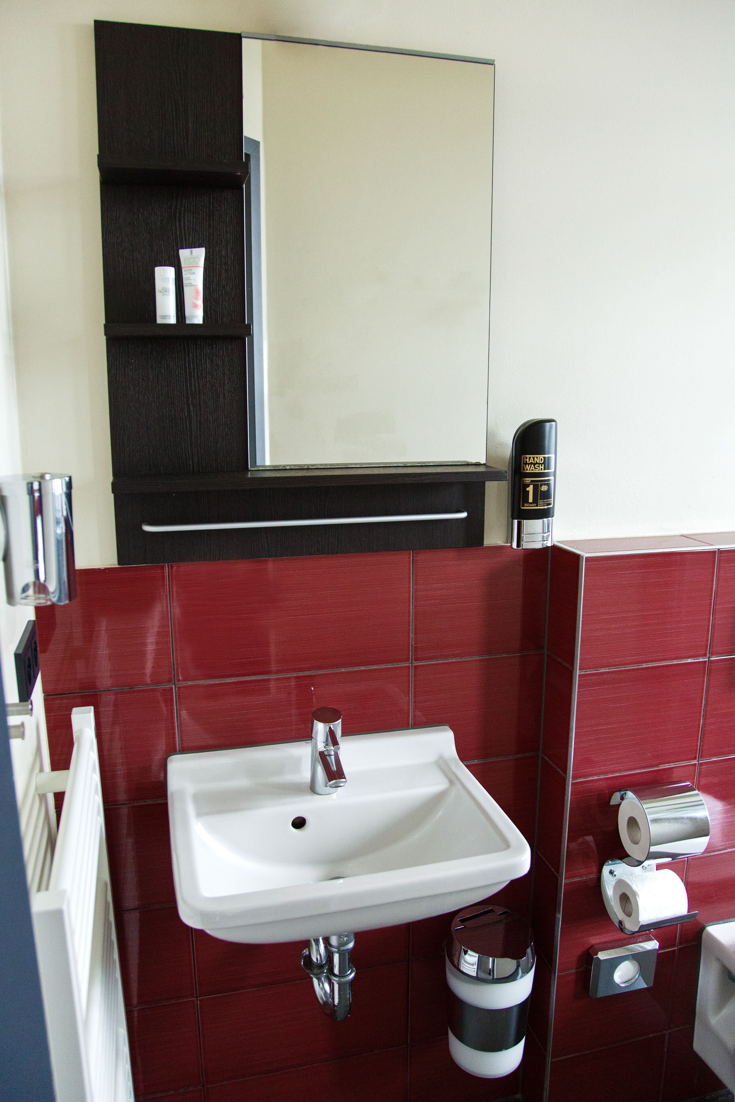 Un baño con lavamanos blanco, baldosas rojas, un espejo, dispensador de gel antibacterial y un cubo de basura.