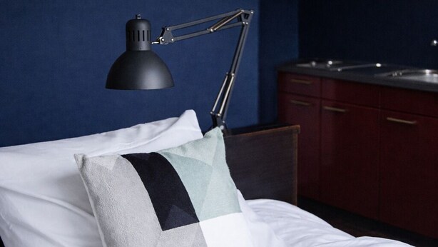 Un lit bien fait, avec un oreiller, une lampe de bureau noire et un mur bleu foncé.