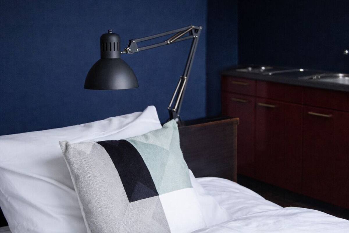 A neatly made bed with a pillow, a black desk lamp, and a dark blue wall.