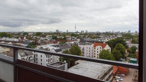 Une vue panoramique sur une ville, prise depuis un point d’observation élevé, avec un mélange de bâtiments résidentiels et commerciaux, une tour imposante et un ciel nuageux.