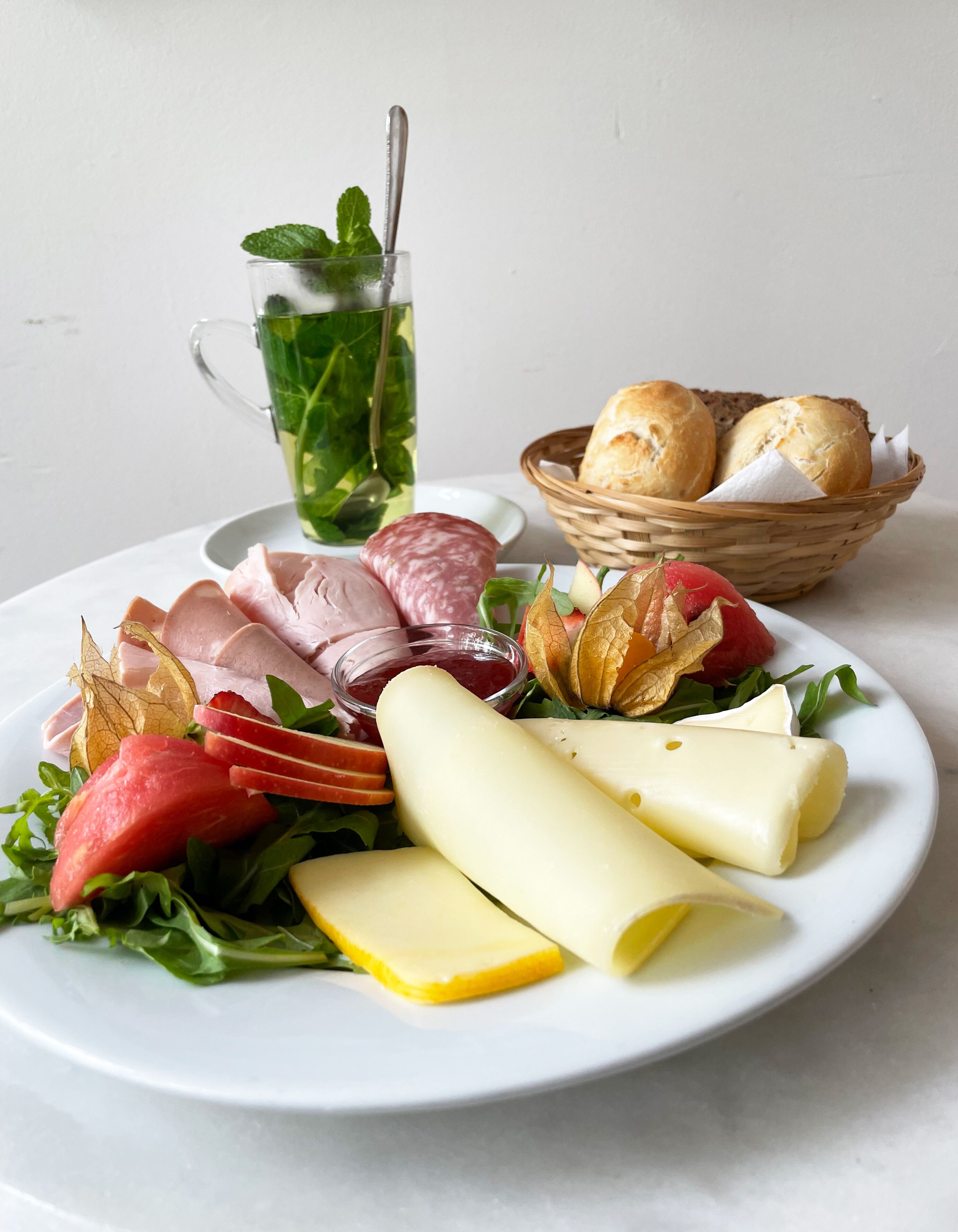 Une assiette garnie d’un assortiment de fromages, de charcuteries et de légumes, avec un panier de pain et un verre de thé glacé en arrière-plan.