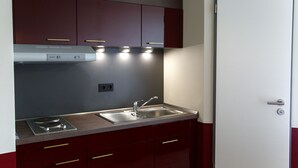 Une cuisine compacte dotée d’un évier en acier inoxydable, d’une hotte intégrée et de meubles de couleur rouge foncé avec des poignées dorées.