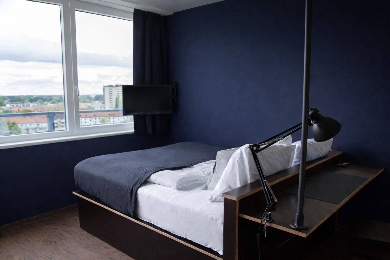 Una camera da letto moderna con un letto, una scrivania, una sedia, una lampada da scrivania e una TV appesa al muro.