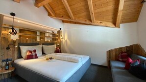 A modern hotel room with a wooden ceiling, a large bed with white linens, a bedside table with a bottle, and a built-in bench with cushions. - Hotel Jagerhof (Kaunertal)