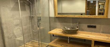Un baño moderno con ducha de vidrio, tocador de madera con lavabo y un espejo grande con marco de madera.